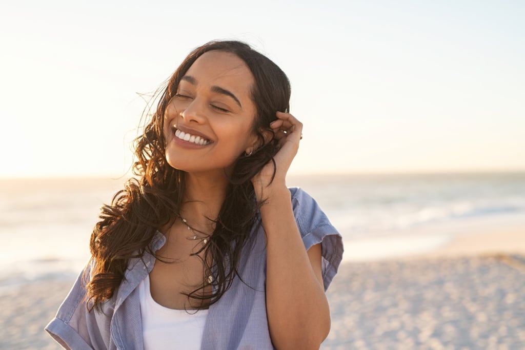 Person smiling with eyes closed, standing on a sunny beach.